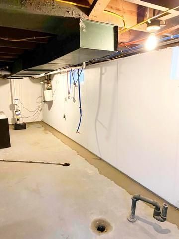 Basement utility area with exposed ceiling beams, white wall, hanging wires, and concrete floor.