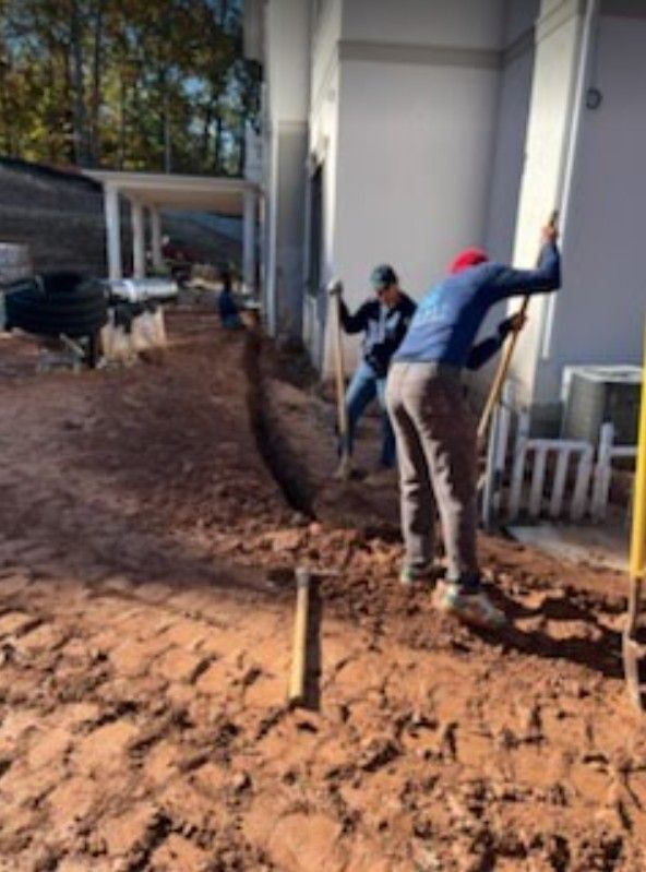 Workers shoveling mud beside a building, with a muddy ground and trees in the background