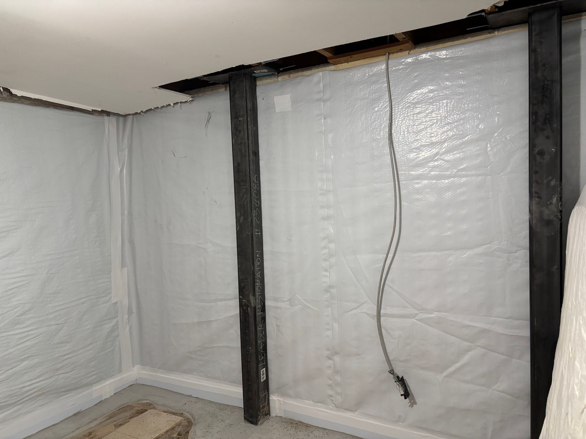 Plastic-covered basement wall with vertical support posts and a hanging electrical cord