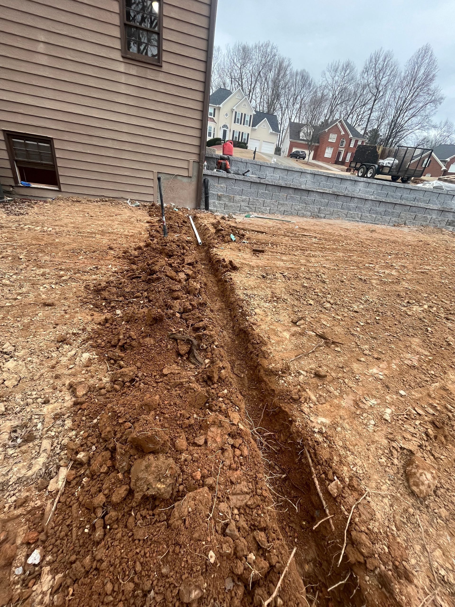 A muddy trench cuts through a leaf-covered yard beside a brown house on a cloudy day.