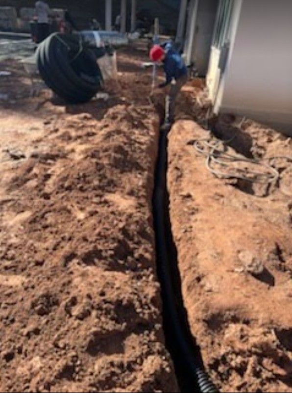 Trench dug alongside a house, with two workers in the background and a large pipe on the left.