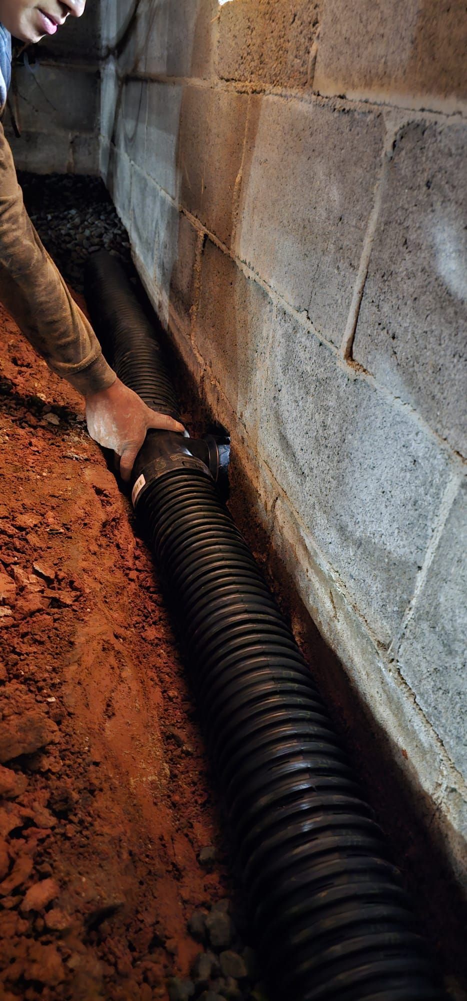Worker installing black corrugated pipe along a dirt trench beside a concrete wall