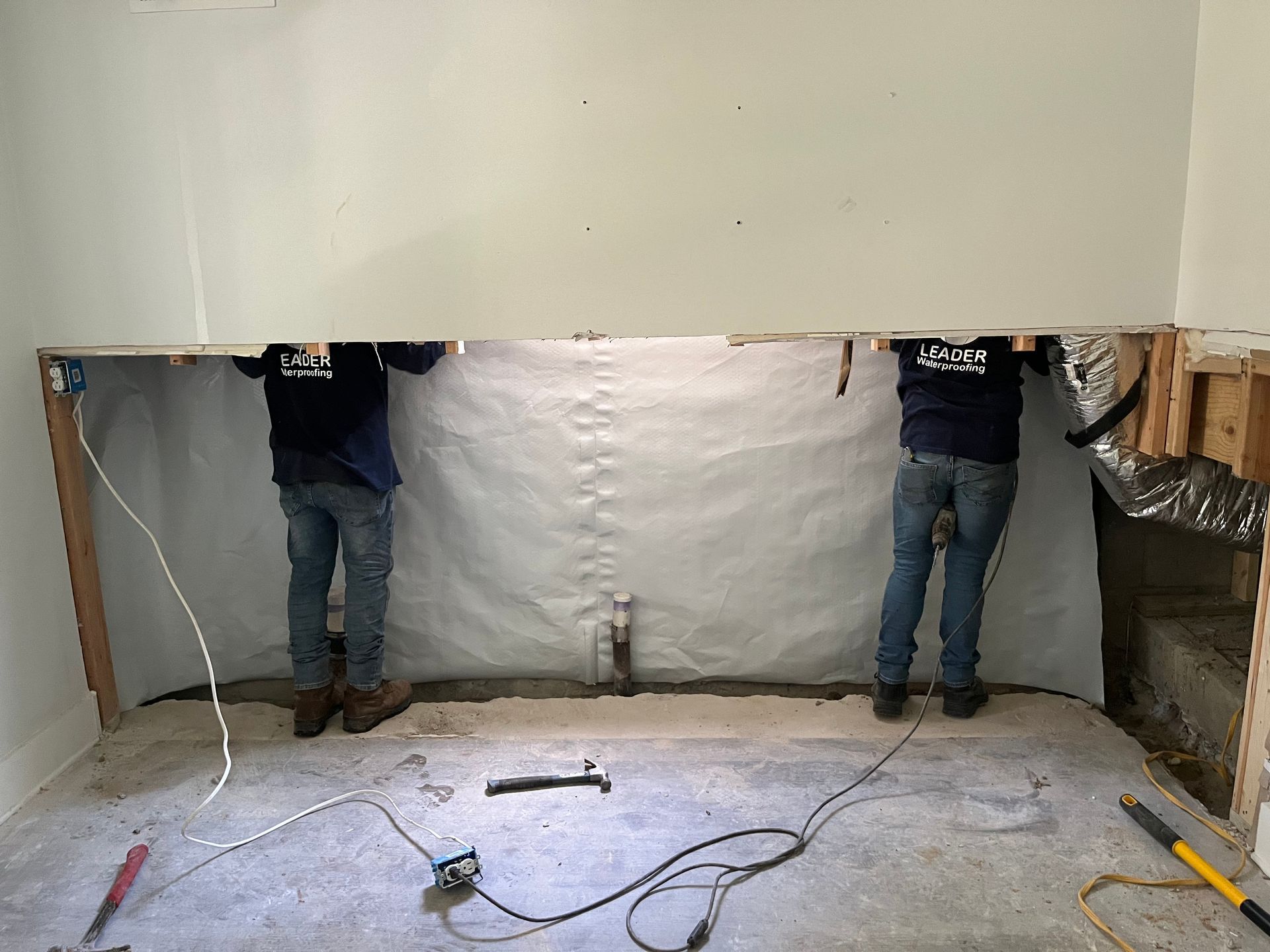 Two workers standing in a partially demolished room with exposed walls, wires, and construction tools on the floor