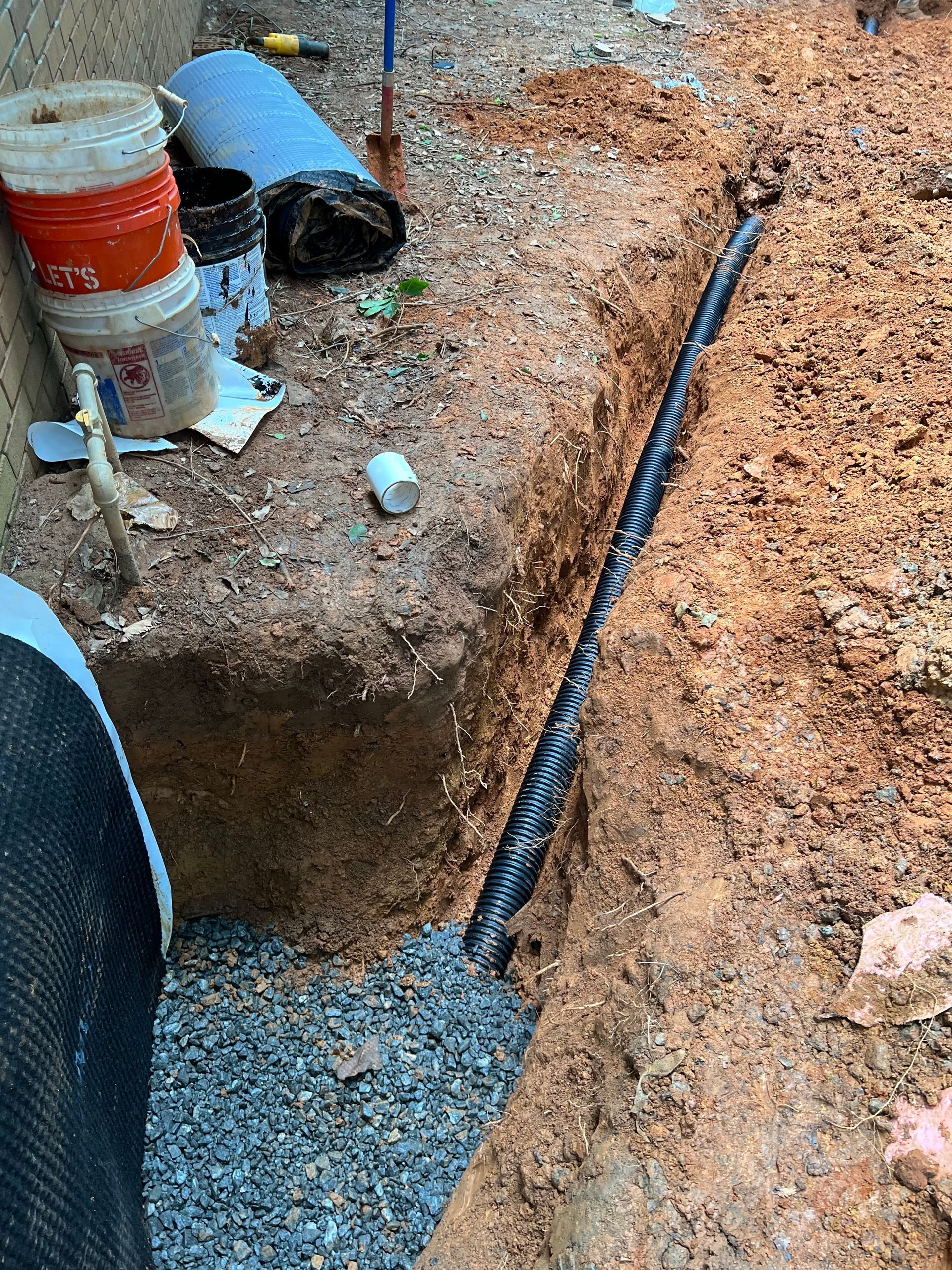 Construction trench with gravel, orange buckets, and a black pipe laid along a dirt edge