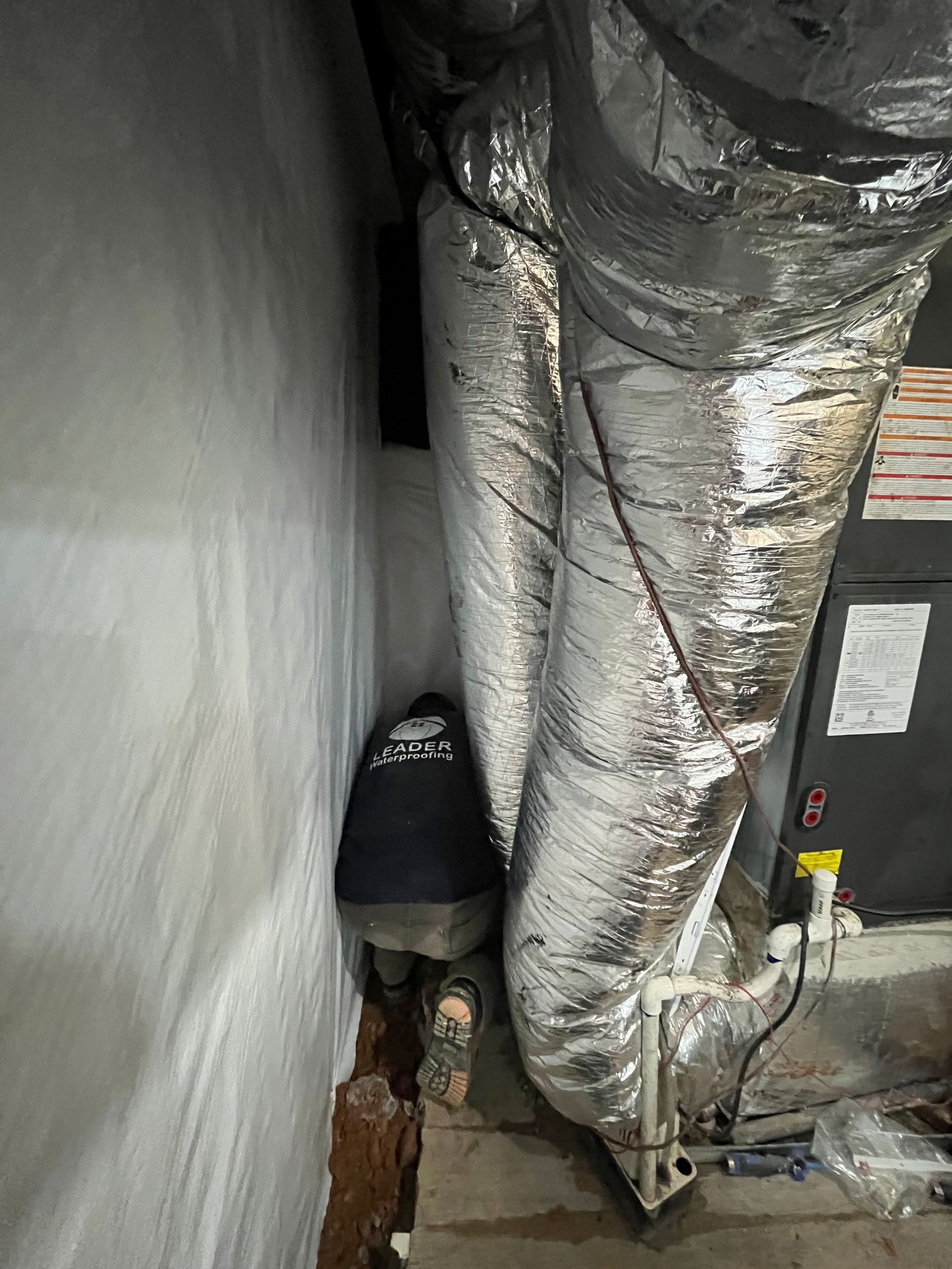 Person crouching in a narrow utility space beside large insulated ducts and HVAC equipment