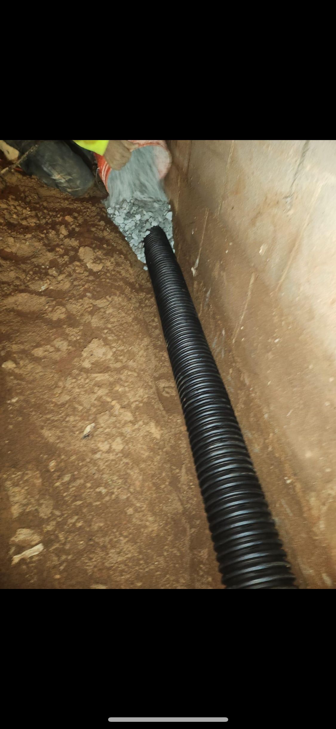 Black corrugated pipe entering a dirt trench beside a concrete wall, wrapped in gray insulation at the joint