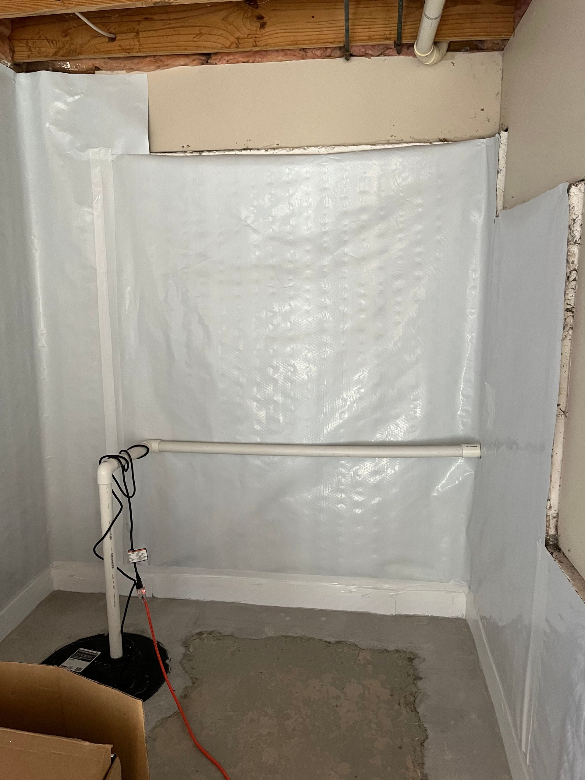 Basement wall wrapped in plastic sheeting with a pipe and unfinished concrete floor