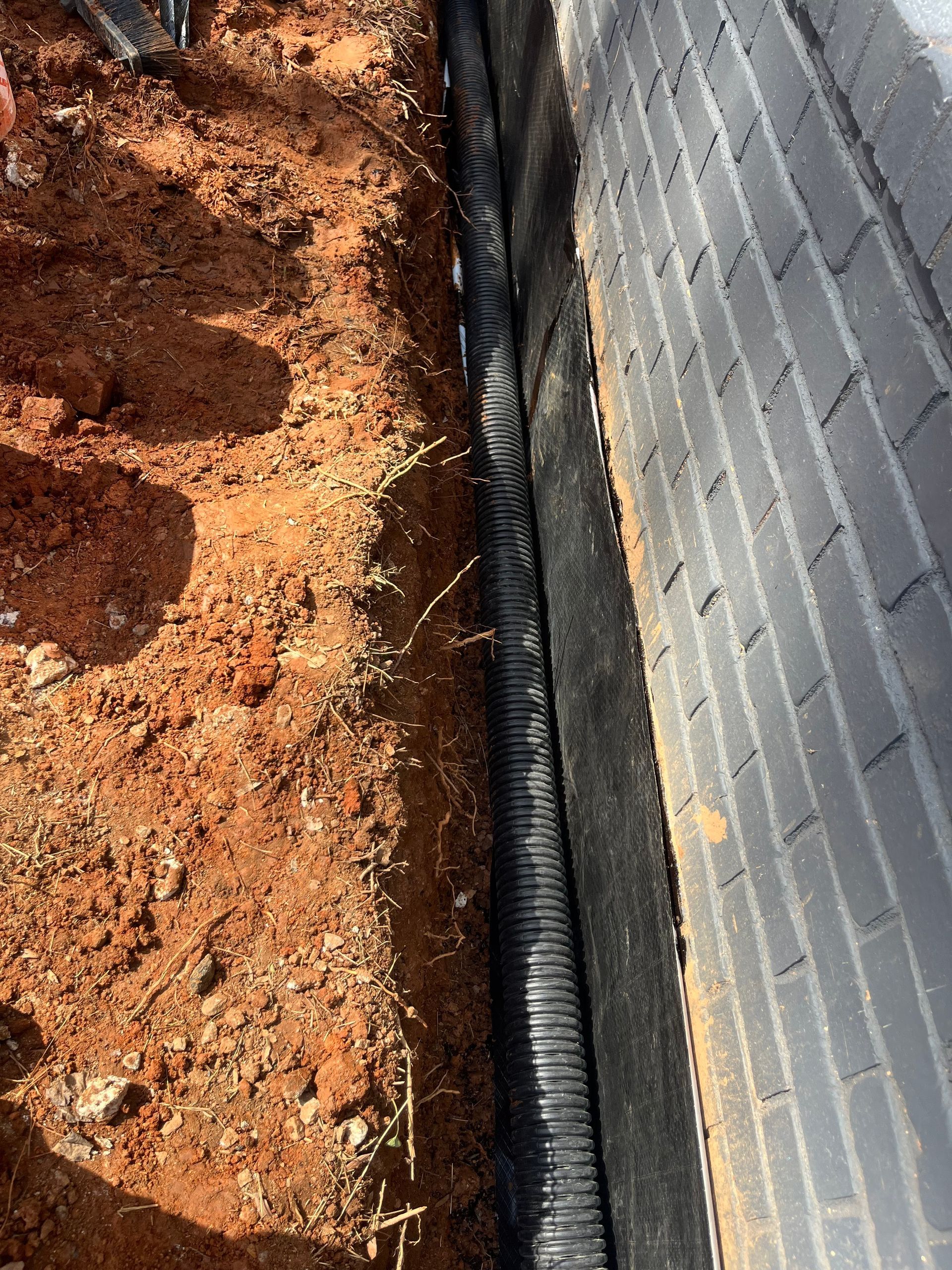 Dirt trench beside a gray brick wall with a black drainage pipe along the ground