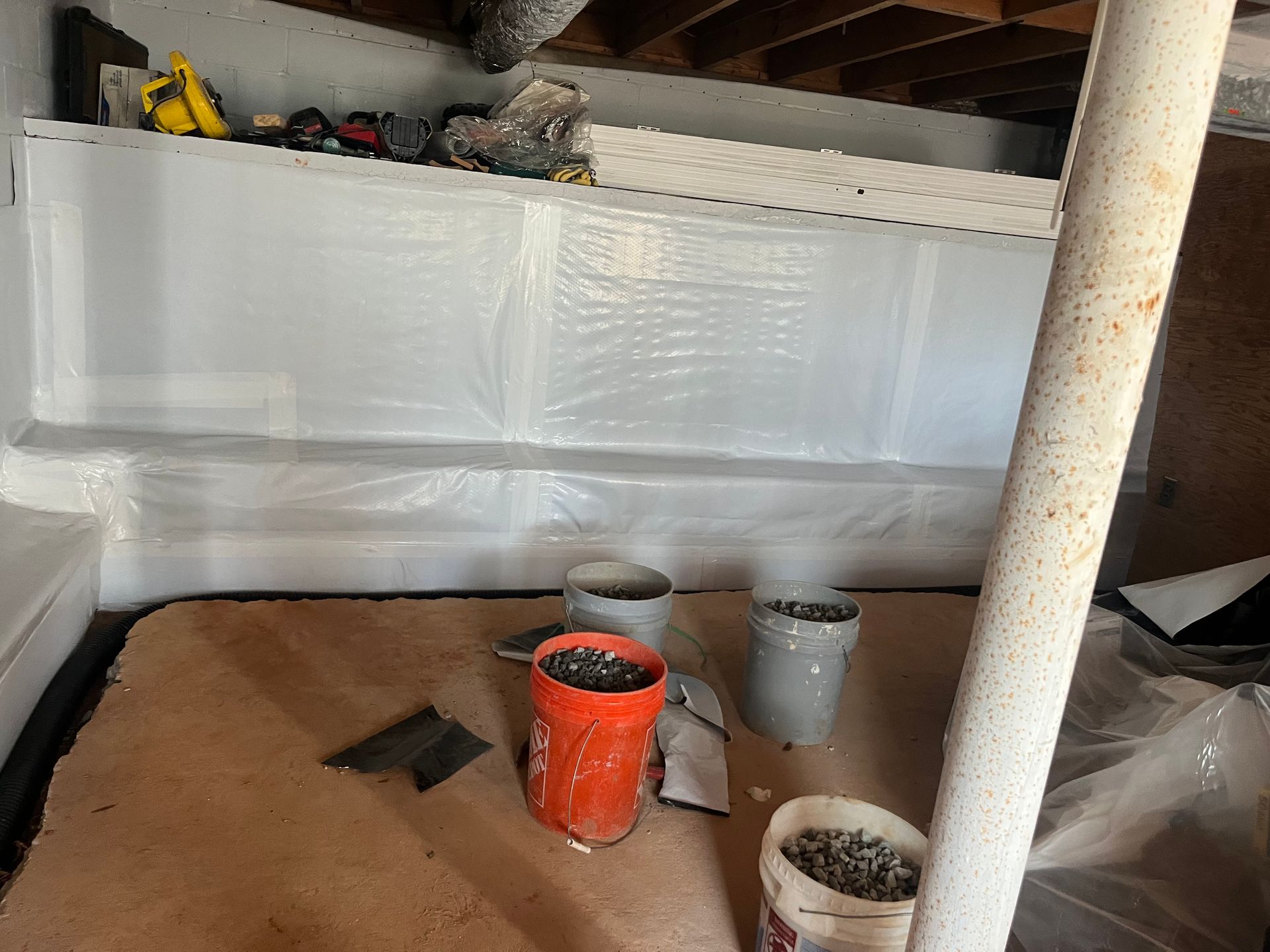 Basement work area with buckets, tools, and plastic-covered walls under exposed beams