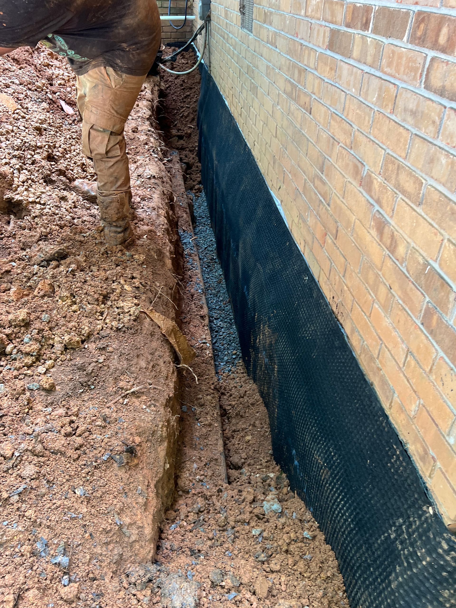 Excavated trench beside a brick wall, lined with black waterproofing mesh and muddy soil.