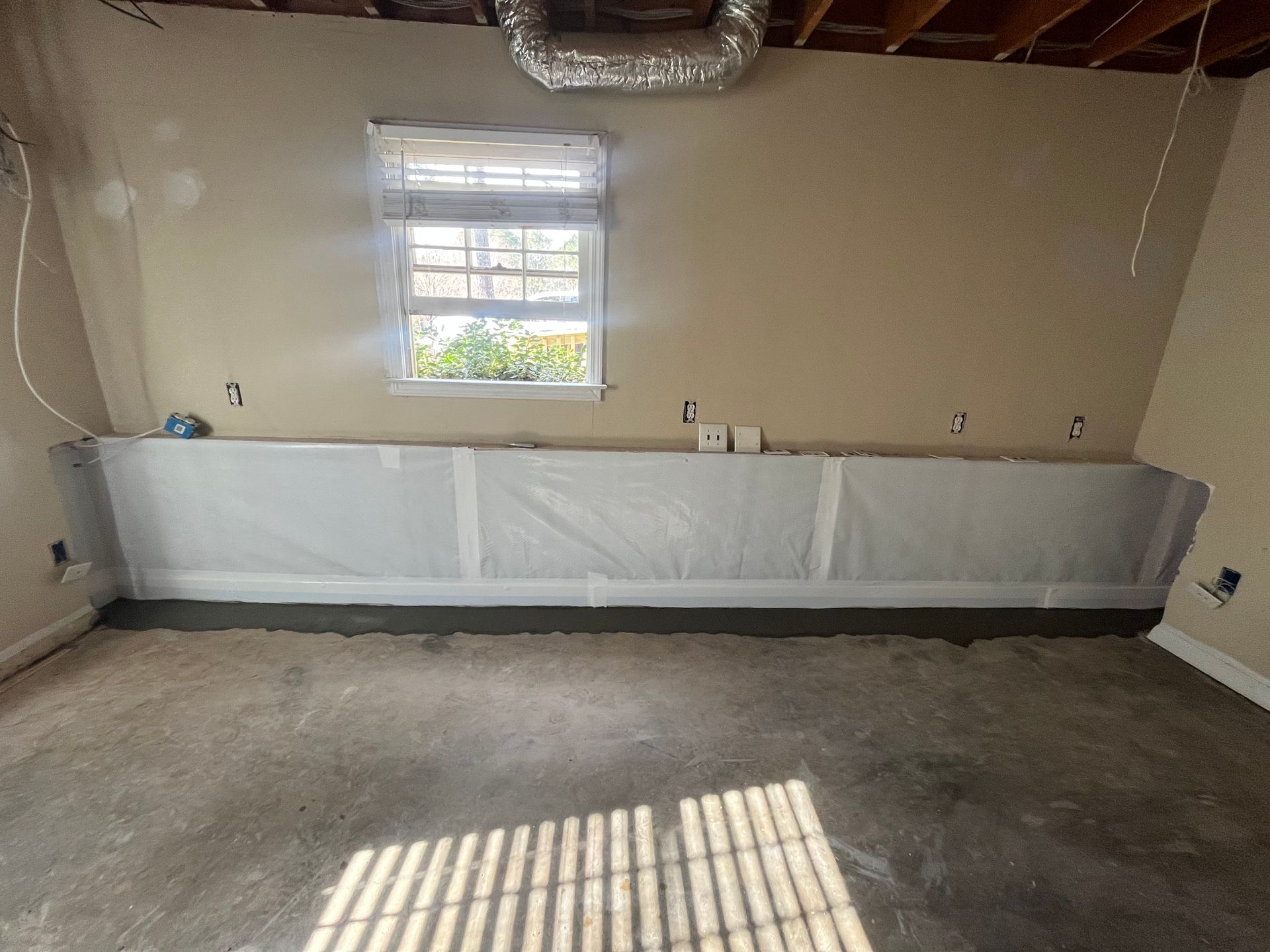 Empty unfinished room with beige walls, concrete floor, small window, and covered baseboards near ceiling beams.