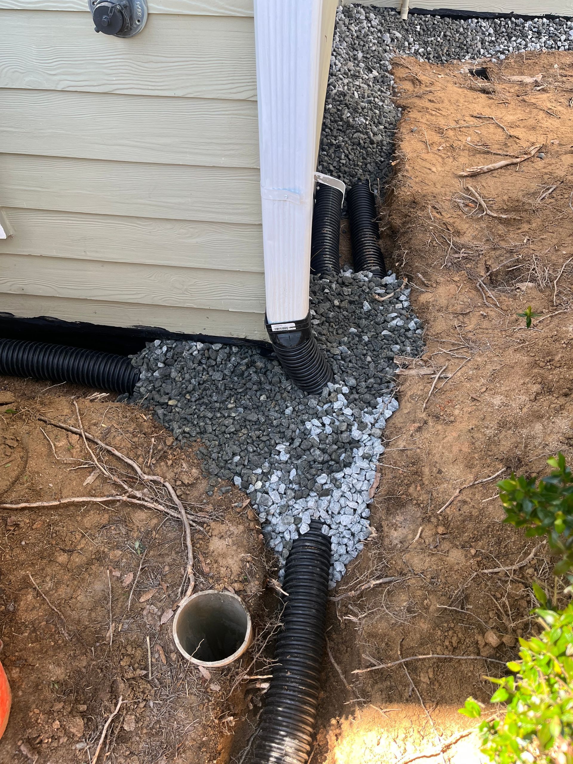 House foundation with black drainage pipe and gravel trench beside tan siding and dirt yard
