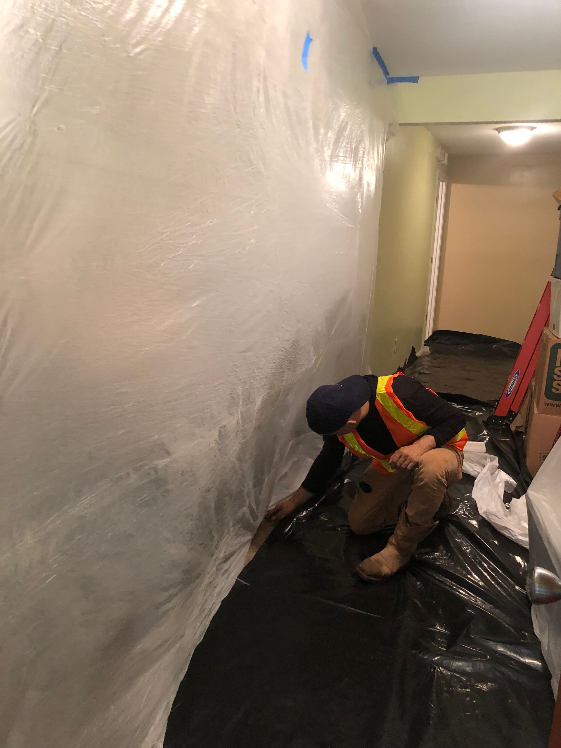 Worker crouching in a hallway, applying a black protective floor covering beside a wall.