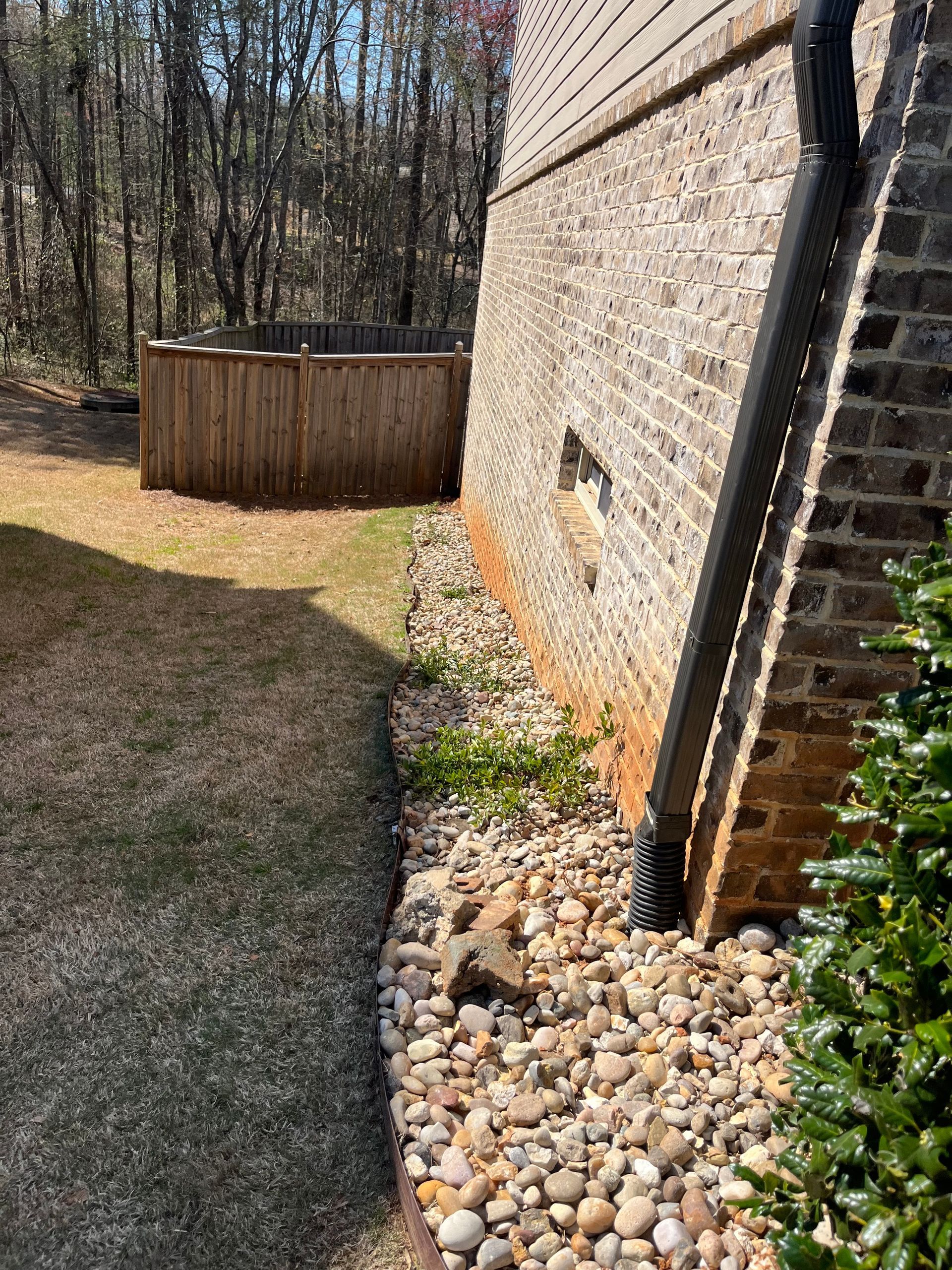 Exterior house wall with stone bed, narrow gravel strip, and curved lawn beside a wooded yard