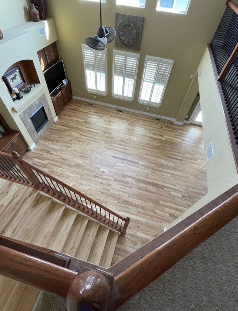 View from a staircase overlooking a living room with wooden floors, fireplace, and large windows.