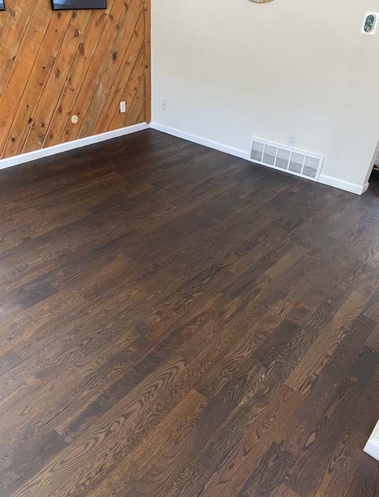Dark wood floor in a room with a wood panel wall and a white wall, white trim and a vent.