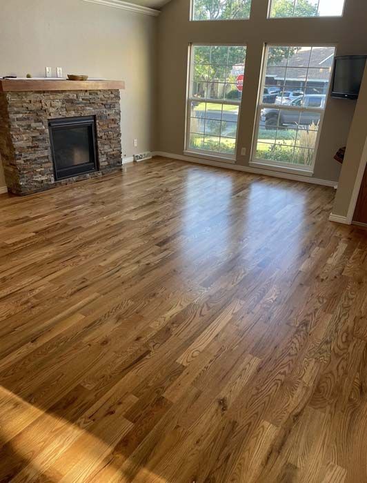 Hardwood floors in a living room with a stone fireplace, large windows, and sunlight.