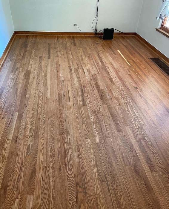 Hardwood floor in a room, light brown with reddish tones. Baseboard, wall and trim visible.