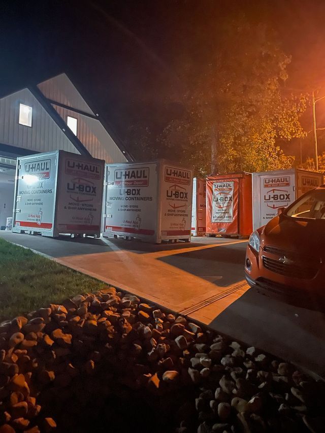 Moving trucks parked in a driveway beside a house at night, lit by orange streetlights.