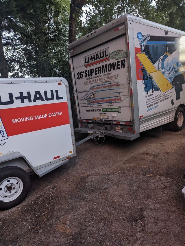 Two U-Haul moving trucks parked on a dirt lot near trees, one labeled 26' SuperMover.