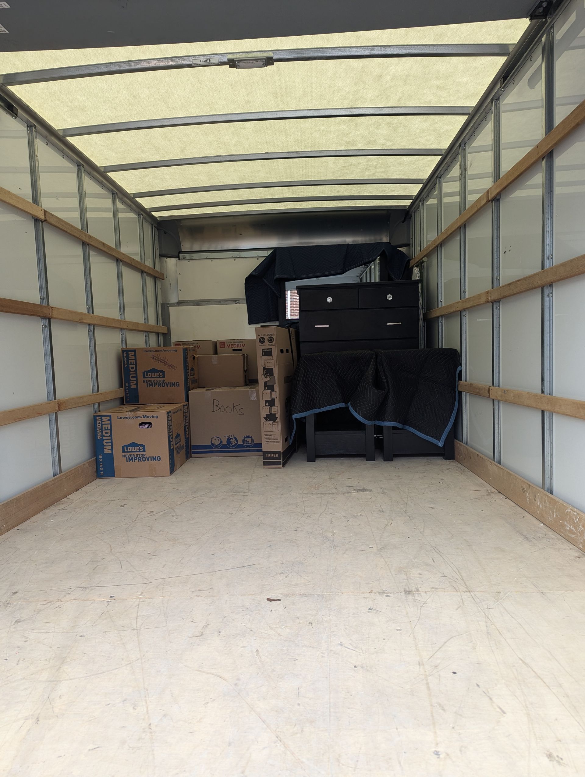 Inside an open trailer, stacked cardboard boxes sit beside a large black machine on the floor.