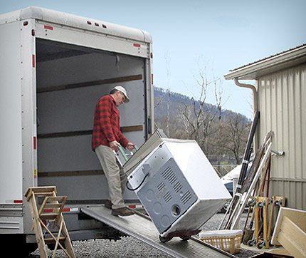 Man loading a tilted washing machine down a ramp from a moving truck in a driveway