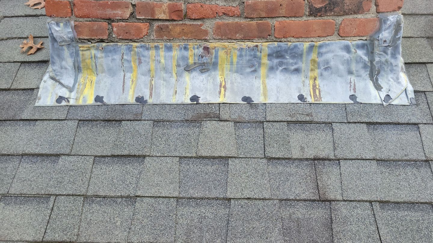 Close-up of a chimney flashing on a shingled roof with yellow sealant streaks.