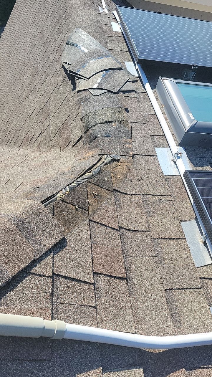 A roof ridge showing damaged, torn asphalt shingles and exposed roof deck near solar panels and a skylight.