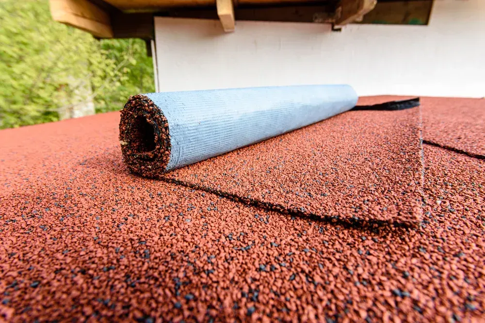 Roll of roofing material on a red shingled roof, under a white wall and wooden structure.