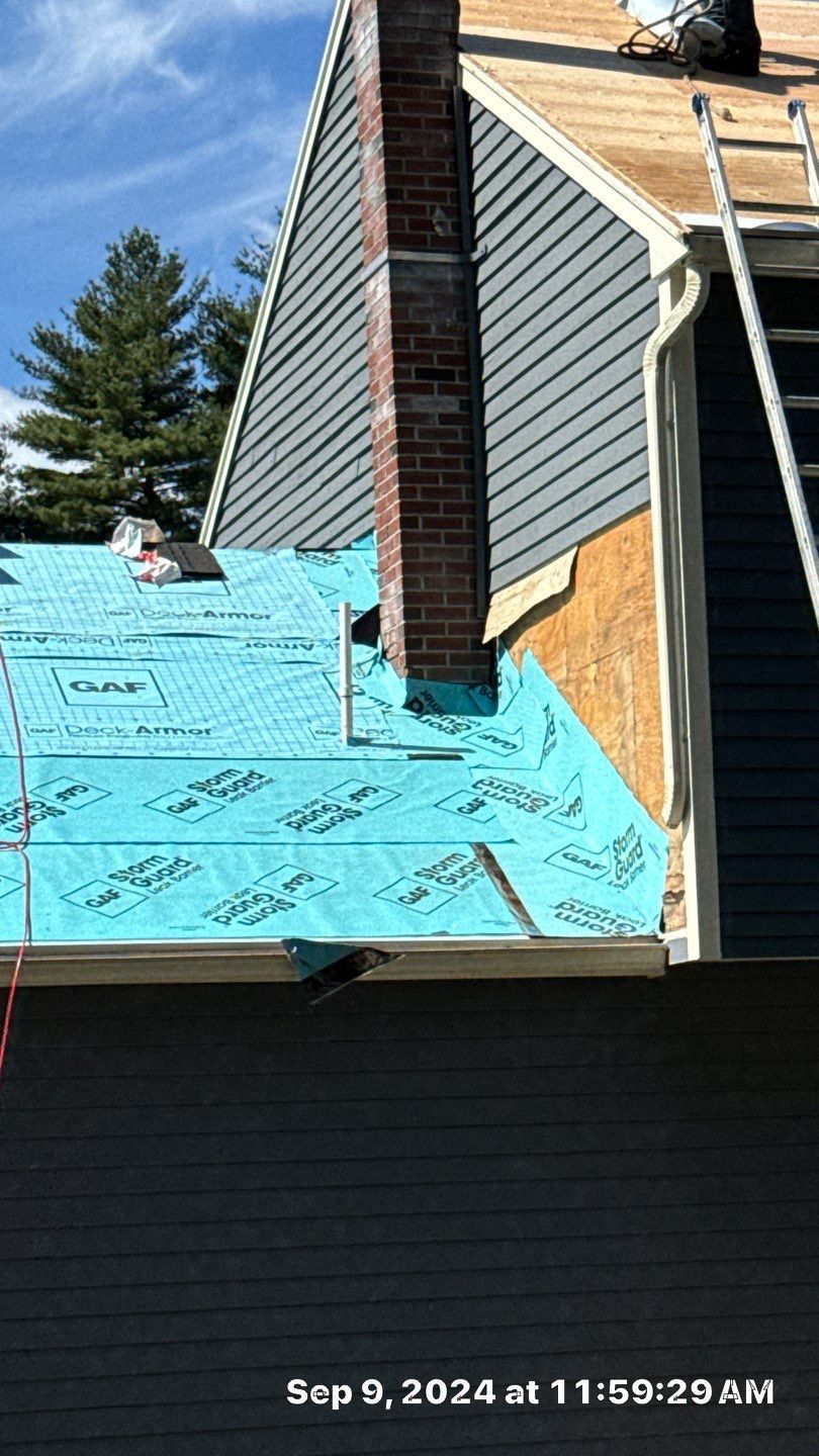 A roof under construction featuring blue GAF underlayment, a brick chimney, gray siding, and a ladder against the eaves.