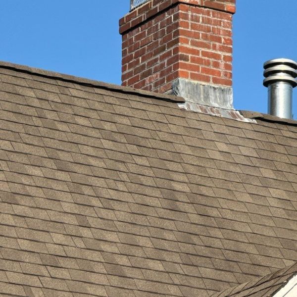 Brown shingled roof with a red brick chimney and a silver vent against a blue sky.