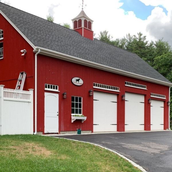 Red barn with three white garage doors, white fence, and black asphalt driveway.