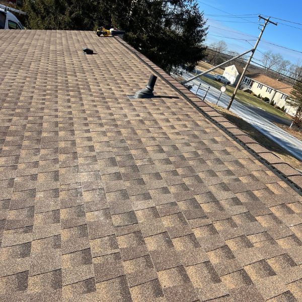 Brown asphalt shingle roof on a sunny day with a black vent pipe and power lines in the background.