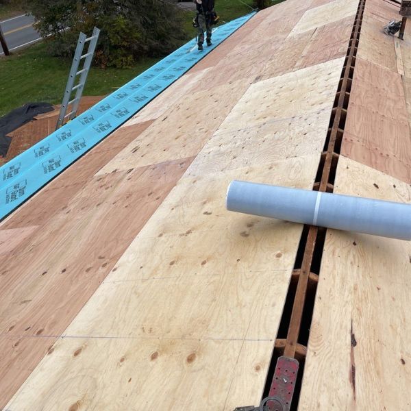 Rooftop with plywood, a long channel, and a roll of gray material. A worker is in the distance.