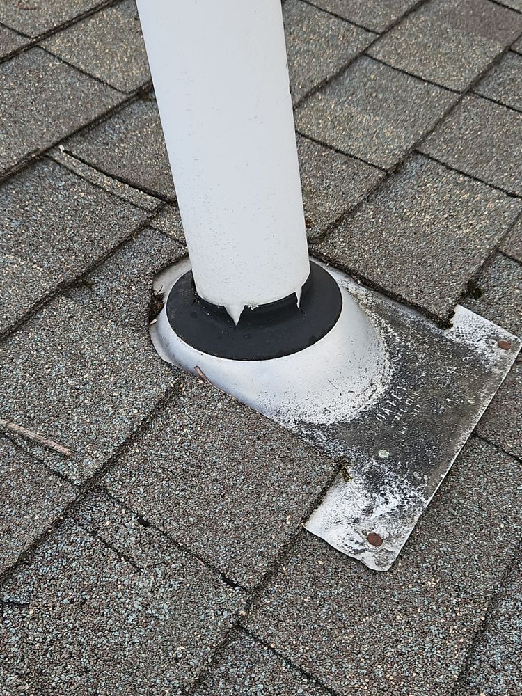 White pipe emerging from roof shingles with a metal flashing and black rubber seal.