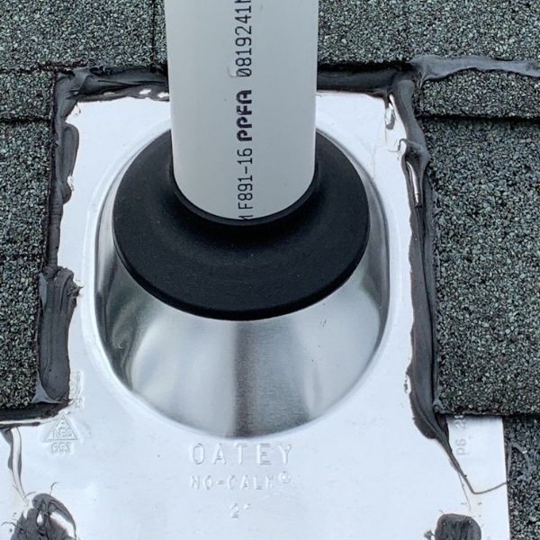 A metal plumbing roof boot flashing sealed with gray caulk around a white PVC pipe on gray asphalt shingles.