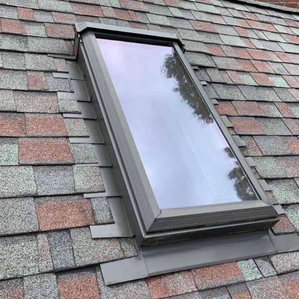 A rectangular skylight installed on a sloped roof with multi-colored gray and reddish-brown asphalt shingles.