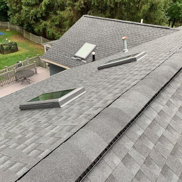 An overhead view of a gray shingled residential roof with three skylights, a roof vent, and a yard visible in the background.