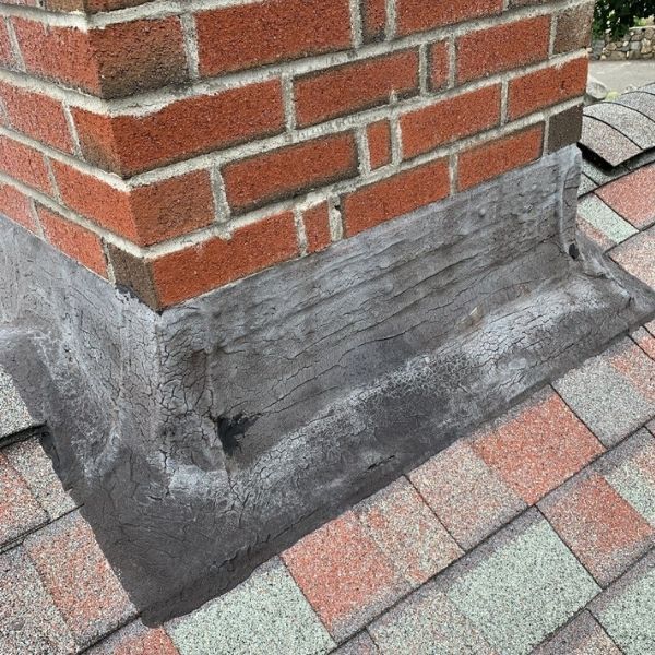 A brick chimney on a shingled roof with deteriorating, patch-repaired flashing at the base.