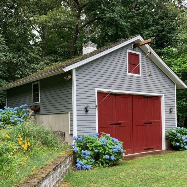 A grey garage with a peaked roof, bright red double doors, and a small loft window, surrounded by hydrangea bushes.