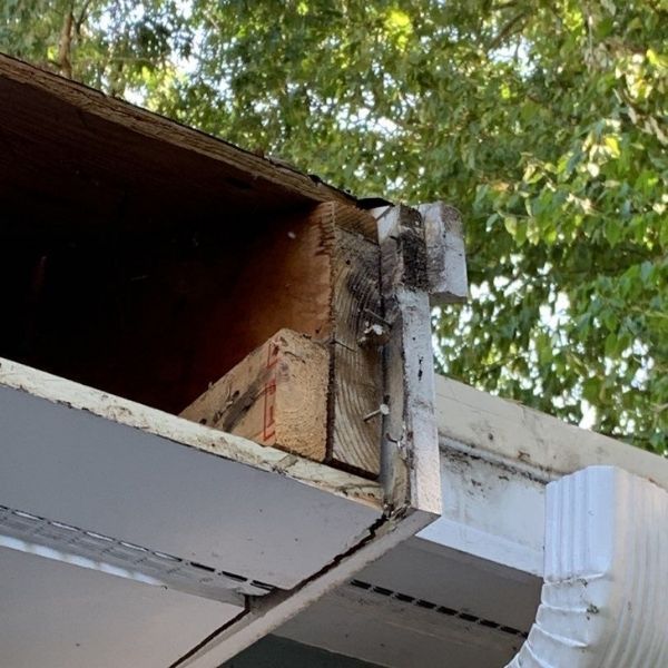 A close-up view of an open, damaged roof soffit showing exposed wooden framing and rusted hardware near a downspout.