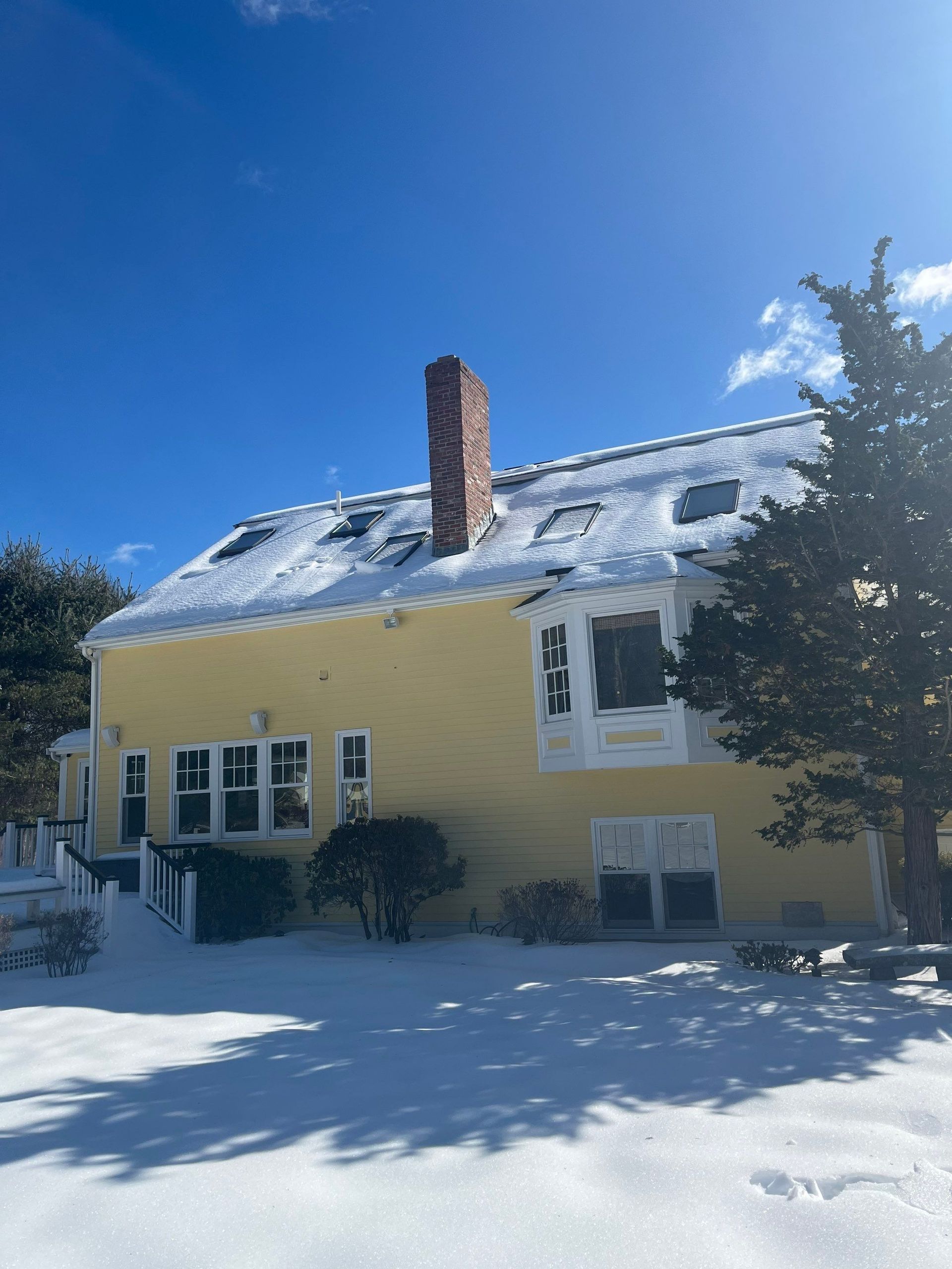 Yellow house with snow-covered roof, chimney, and surrounding ground; clear blue sky.