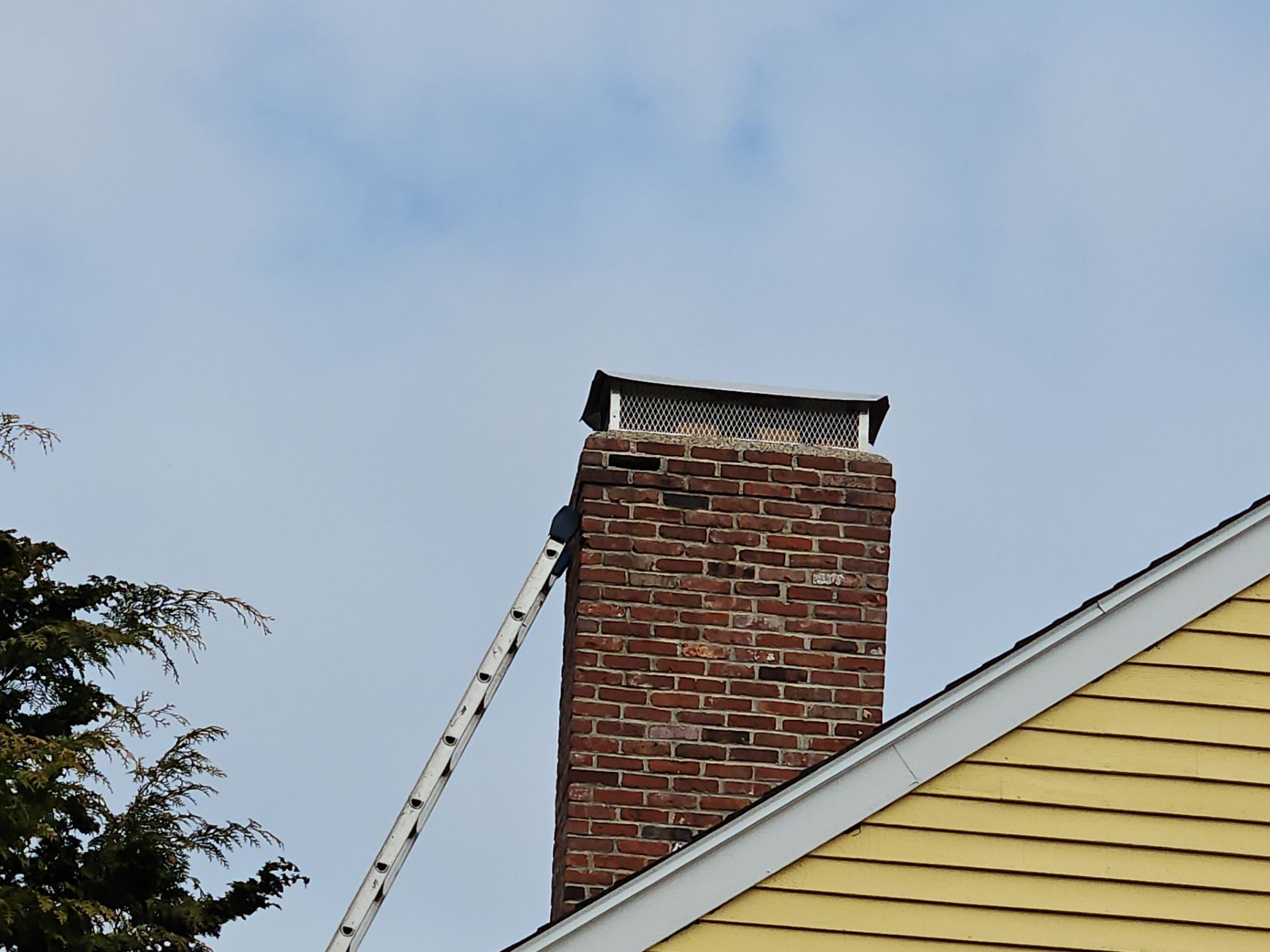 Brick chimney with attached ladder, topped by a gray cap, next to a yellow house with a blue sky background.