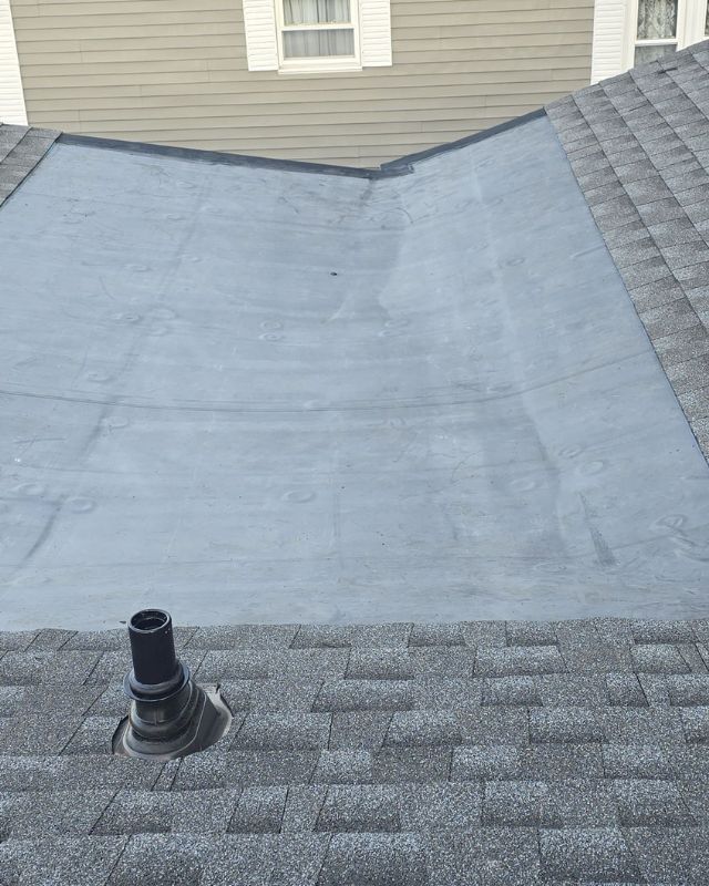 Gray shingled roof with a gray flat section; black vent pipe and window in background.