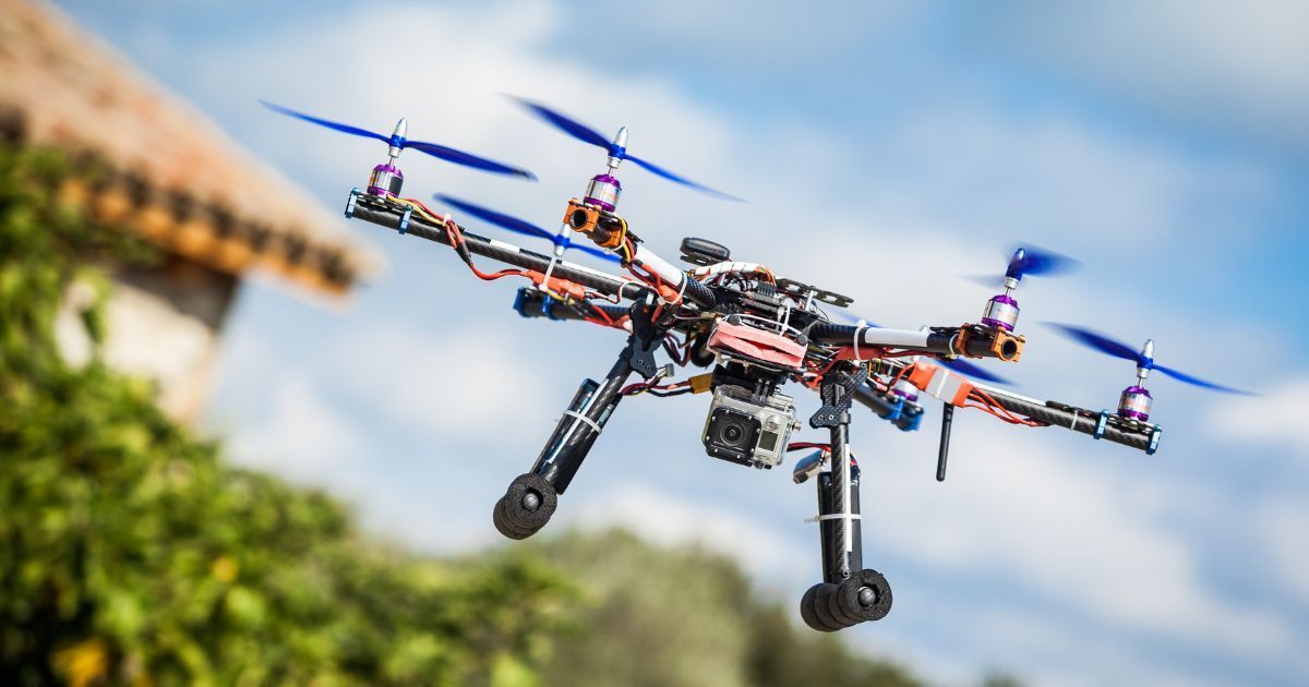 Drone in flight, blue propellers, camera mounted, blurred background of a building and greenery.