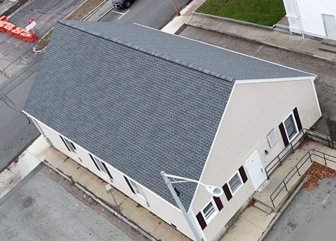 Aerial view of a building with a dark gray roof and beige siding. The building has several windows and a ramp.