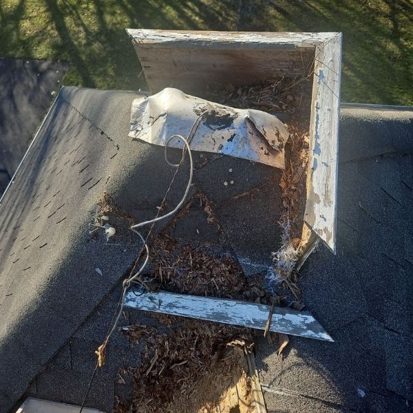 Damaged roof vent with exposed wood, loose metal flashing, and debris accumulating on dark asphalt shingles.