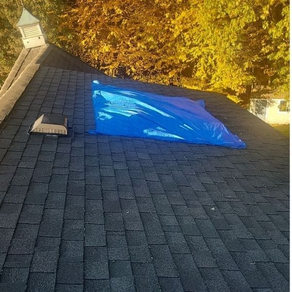 A blue plastic tarp covers a section of a dark-shingled roof next to a vent, suggesting a temporary repair.