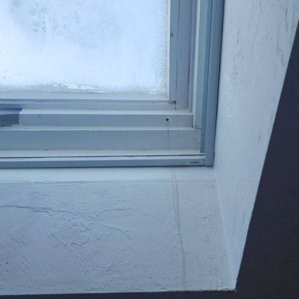 A close-up view of a white window frame set in a textured wall, showing a thin vertical crack in the paint below the sill.