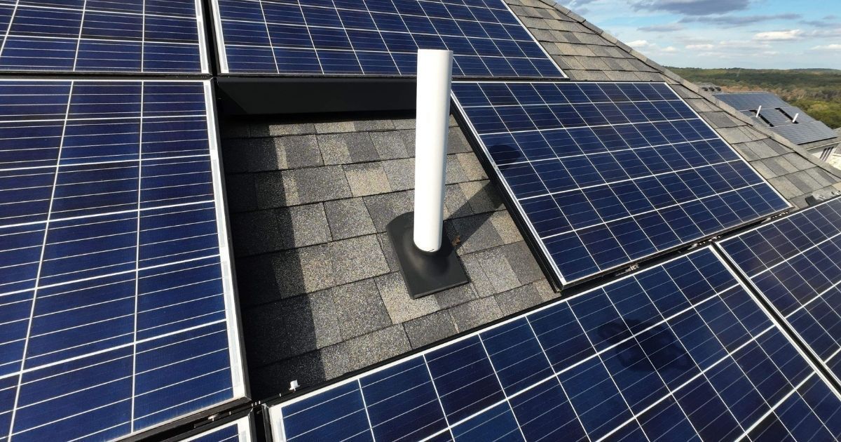 A white plumbing vent pipe surrounded by a grid of dark solar panels on a residential asphalt shingle roof.